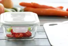 Glass food container with sliced tomatoes sits near carrots and fresh produce