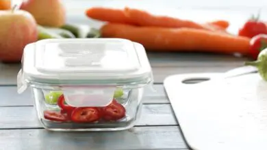 Glass food container with sliced tomatoes sits near carrots and fresh produce