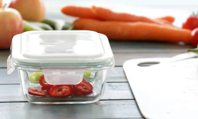 Glass food container with sliced tomatoes sits near carrots and fresh produce