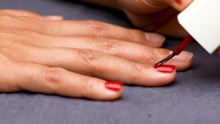 A brush applies red polish to short nails in a close-up manicure