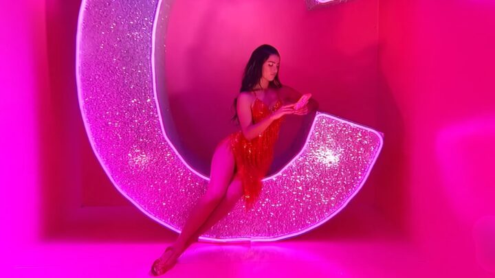 Charli D’Amelio in a red dress sits on a bright pink set and looks at her phone