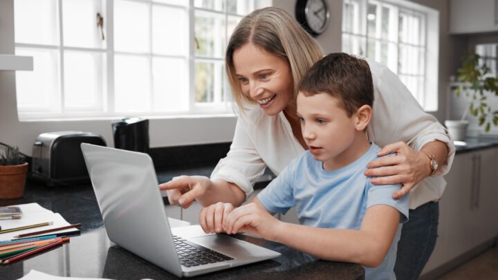 A mother helps her son use a laptop at a kitchen counter