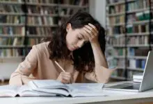Student writes a college essay at a library desk beside open books and a laptop