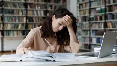 Student writes a college essay at a library desk beside open books and a laptop