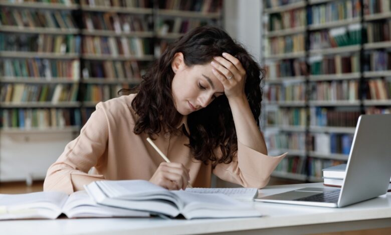 Student writes a college essay at a library desk beside open books and a laptop