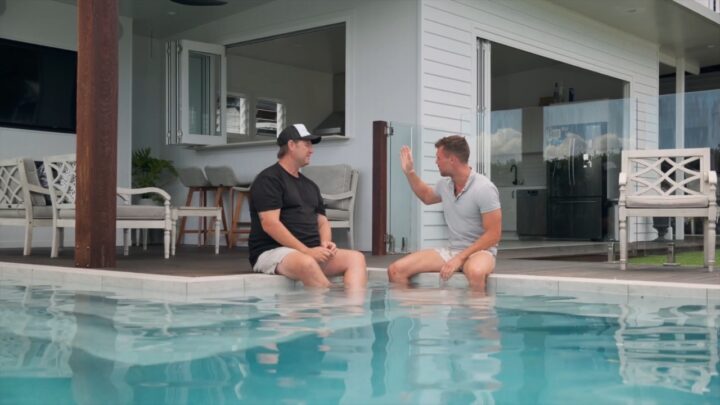 Two men sit at the edge of a backyard pool beside a modern patio area